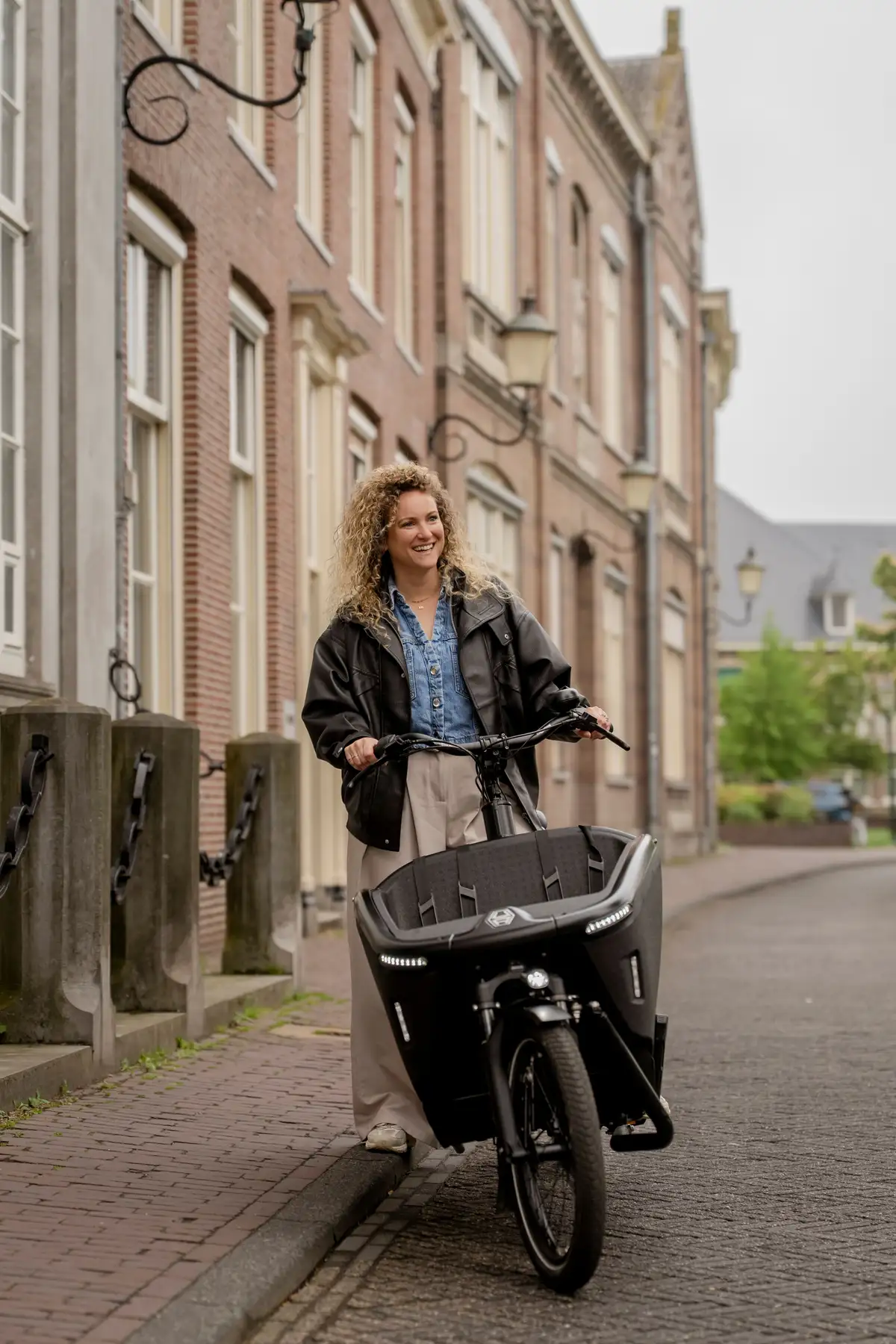 Vrouw staat lachend naast een zwarte elektrische bakfiets in een stadsstraat.