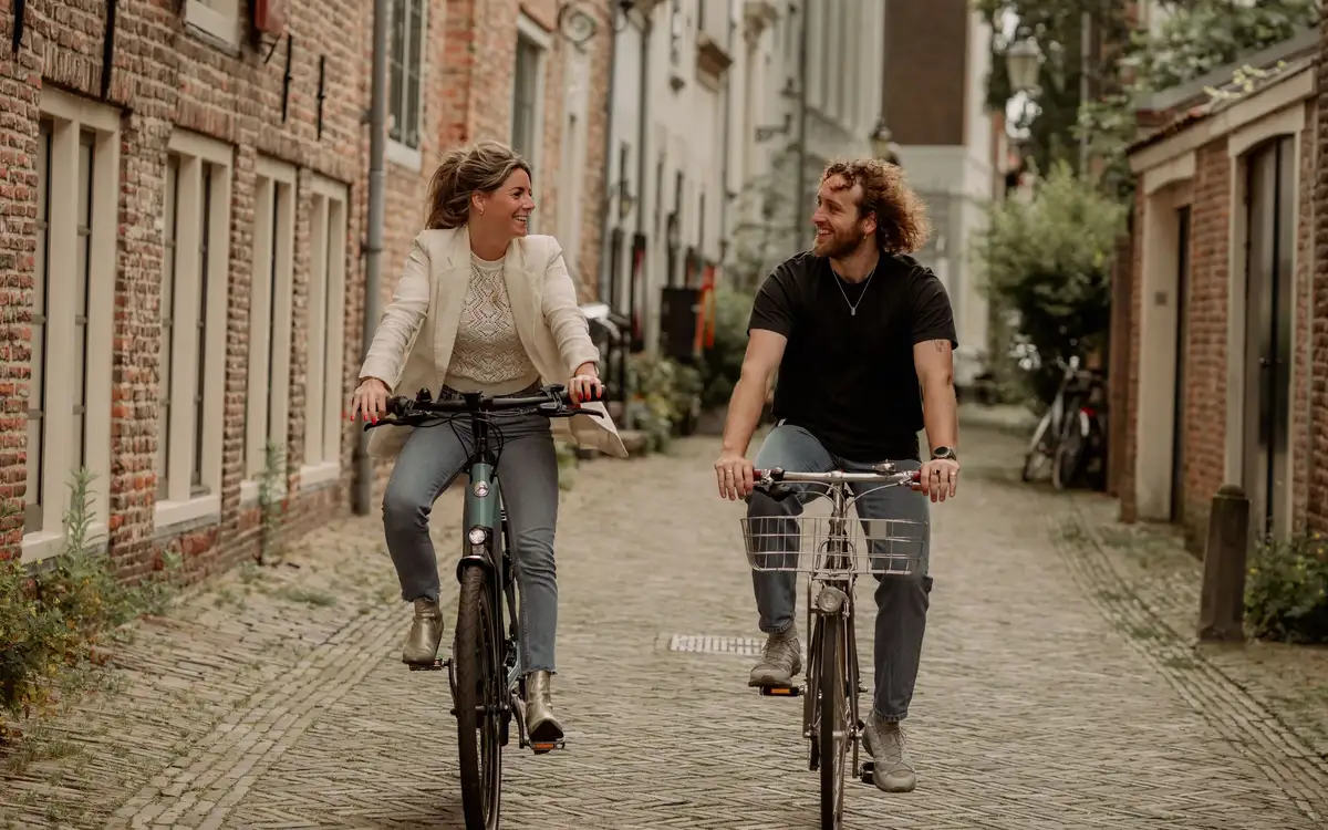 Twee collega's fietsen lachend naast elkaar op een leasefiets door een stadsstraat.